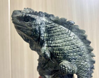 Puede incluir: Una escultura detallada de un lagarto, tallada en piedra con tonos grises y verdes. El lagarto está posado en una posición dinámica, con intrincados detalles de escamas y textura. La escultura es un objeto decorativo.