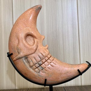 May include: A carved orange crescent moon sculpture with a skull face, displayed on a black metal stand.