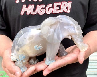 May include: A carved stone elephant figurine in shades of grey and white, with blue-green accents. The elephant is held in a person's hands. The shirt in the background says "I'M A HUGGER!"
