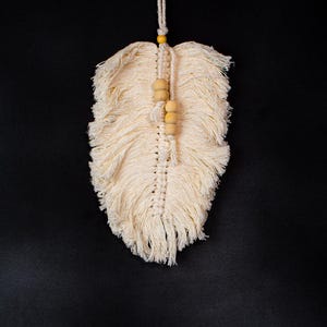 May include: Cream-colored macrame feather wall hanging with wooden beads. The feather design is made of knotted cotton cord, with a fringe at the bottom. A small yellow bead is at the top, with a cream-colored cord for hanging.