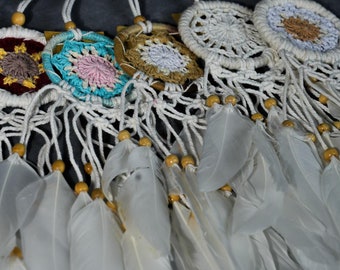 May include: Four white dreamcatchers with colorful woven designs and white feathers hanging from them. The dreamcatchers are strung together with white cord and wooden beads.