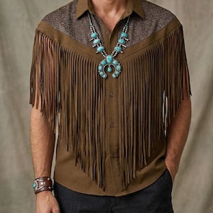 Puede incluir: Camisa marrón con flecos en el pecho y mangas, combinada con pantalones grises oscuros. La camisa tiene un canesú texturizado y se complementa con un collar y pulsera turquesa, con un estilo western.
