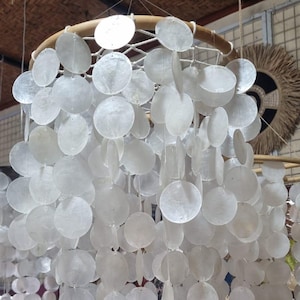 Könnte beinhalten: Ein hängender Kronleuchter aus zahlreichen weißen, runden, muschelartigen Scheiben. Die Scheiben sind an einem Holzring aufgehängt und durch Schnüre verbunden, wodurch ein geschichteter, kaskadierender Effekt entsteht. Für die Inneneinrichtung.