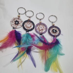 Puede incluir: Cuatro llaveros atrapasueños con aros plateados. Cada atrapasueños tiene un diseño circular tejido en blanco, negro, morado y morado oscuro. Plumas coloridas en tonos rosa, morado, azul y amarillo cuelgan debajo.