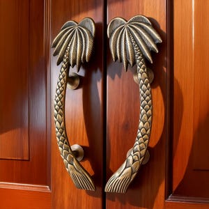 May include: A pair of bronze-colored door handles shaped like palm trees. The handles are mounted on a dark wooden door. The palm fronds are at the top, and the trunks curve down to the base.