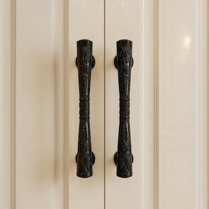 May include: Two black, ornate cabinet pulls with a detailed, textured design. The pulls are vertically oriented and mounted on a cream-colored cabinet door. The design features floral and scrollwork patterns, adding a touch of elegance.