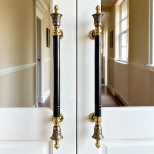 May include: Pair of ornate door handles. Each handle features a black, cylindrical grip with gold-colored decorative elements at the top and bottom. The handles are mounted on a white door, with a hallway visible in the background.
