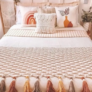 May include: A bed with a cream-colored woven blanket and matching pillows. The pillows feature a rainbow, a macrame design, and a vase with leaves. The blanket has a fringe of colorful tassels.