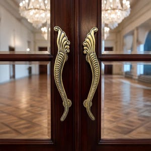 Puede incluir: Primer plano de un par de manijas de puerta de latón ornamentadas en una puerta de madera oscura. Las manijas tienen un diseño curvo en forma de hoja. La puerta es parte de una gran sala con candelabros.