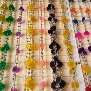 Puede incluir: Filas de guirnaldas decorativas coloridas. Cada hebra presenta una mezcla de piezas iridiscentes en forma de concha y pequeñas cuentas cuadradas de color plateado. Los colores incluyen verde, amarillo, rosa, morado, negro y azul. Las guirnaldas cuelgan de una rejilla blanca.