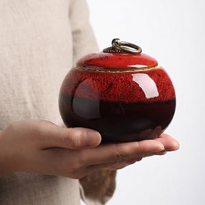 May include: A round ceramic jar with a glossy finish, featuring a vibrant red top that transitions into a deep dark brown base. The lid is topped with a small, circular bronze-colored metal ring handle, providing a functional accent to the container.