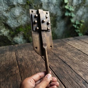May include: A rectangular antique brass door bolt featuring a sliding mechanism with a central handle and a decorative plate. The metal surface has a dark, weathered patina with visible screw holes at the top and bottom for mounting purposes.