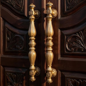 Puede incluir: Primer plano de un par de manijas de puerta doradas y ornamentadas. Las manijas, de unos 30 cm de largo, presentan un diseño detallado en forma de hoja. Están montadas en una puerta de madera marrón oscuro con patrones florales tallados.