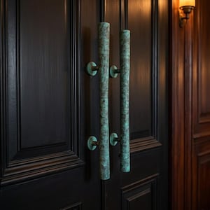 May include: Two vertical door handles with a weathered teal and copper patina finish, featuring intricate etched geometric patterns along their cylindrical length, mounted on a dark brown wooden double door with recessed paneling details.