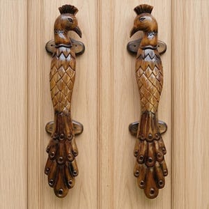 Könnte beinhalten: Zwei Türgriffe aus Holz in Form von Pfauen. Die Griffe sind braun mit detaillierten Feder- und Kopfverzierungen. Jeder Griff hat eine dekorative Montageplatte. Sie sind an einer hellbraunen Holztür befestigt.