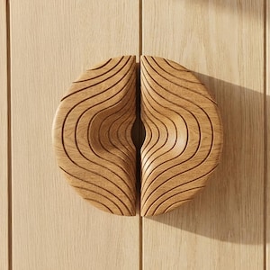 Può includere: Una coppia di maniglie per armadietti semicircolari in legno marrone chiaro con una texture intagliata ondulata e increspata. I due pezzi sono posizionati insieme per formare una forma circolare, mostrando una venatura del legno naturale.