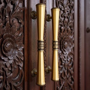 May include: Close-up of two gold-colored door handles with a decorative design. The handles are mounted on a dark brown wooden door with intricate floral carvings. The handles have a cylindrical shape with decorative bands.