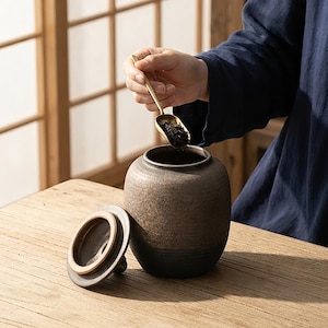 May include: A dark brown ceramic tea canister with a textured, speckled finish and a matching lid placed beside it. A small, slender gold-toned metal scoop is held above the open canister, containing a portion of loose-leaf black tea leaves.