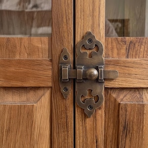 Op de afbeelding: Close-up van een houten kastdeur met een decoratieve antieke messing sluiting. De sluiting heeft een sierlijk ontwerp met een ronde knop en is vastgezet met schroeven. De kastdeuren zijn gemaakt van lichtbruin hout.
