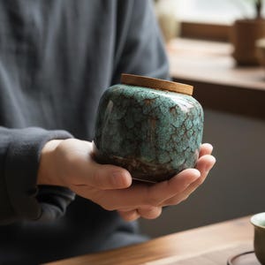 Puede incluir: Un pequeño tarro de cerámica con un esmalte texturizado, verde azulado y marrón. El tarro tiene forma redondeada y una tapa de madera. El tarro está sostenido en las manos de una persona. El tarro es probablemente para guardar té u otros objetos pequeños.