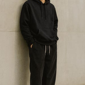 May include: Black hoodie and corduroy pants set. The hoodie has a front pocket and drawstring. The pants have a drawstring waist. The word "BLACK" is printed in white at the top. The outfit is paired with white and beige sneakers.
