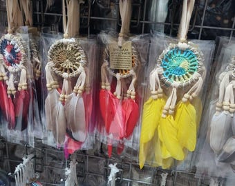 May include: A collection of colorful dream catchers, each with a woven circular center, feathers, and wooden beads. The dream catchers feature various colors, including purple, red, yellow, and gray. They are displayed in clear plastic packaging.