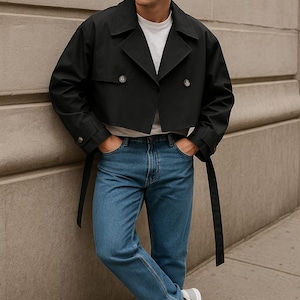 May include: A black cropped trench coat with a double-breasted design, worn with blue jeans and white trainers. The coat has a belt and is over a white t-shirt. The outfit is casual and fashionable.
