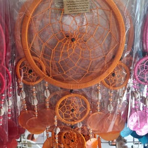 May include: Orange dream catcher with a woven web design, shell accents, and hanging orange shell-shaped pendants. The dream catcher has a tag that reads "Dream Catcher". The dream catcher is hanging in front of a metal grid.