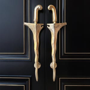 Puede incluir: Un par de manijas de puerta doradas ornamentadas. Las manijas están montadas en una puerta negra con molduras doradas. Las manijas tienen un diseño único y estilizado con una parte superior curva y una forma orgánica texturizada.