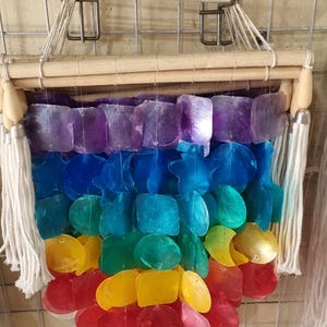 Puede incluir: Un adorno de pared con forma de arcoíris hecho de conchas iridiscentes. Las conchas están dispuestas en un degradado de rojo a morado, con azul, verde y amarillo en el medio. Está sujeto a un marco de madera con borlas blancas.