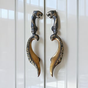 Puede incluir: Par de tiradores de armario de bronce ornamentados. Cada tirador presenta una cabeza de león estilizada en la parte superior y un diseño curvo y decorativo. Los tiradores están montados en una puerta de armario blanca, mostrando sus intrincados detalles y acabado antiguo.