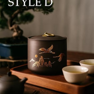 May include: A dark brown tea canister with a gold-coloured lid and handle. The canister features a gold illustration of flying birds, mountains, and a sun. The text "STYLE D" is at the top. The canister sits on a wooden tray with teacups and a teapot.