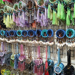 Könnte beinhalten: Eine Wanddisplay mit bunten Traumfänger-Schlüsselanhängern mit Perlen, Muscheln und Quasten. Die Schlüsselanhänger sind in Reihen auf einem Metallgestell angeordnet.