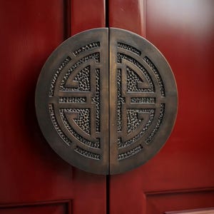 Può includere: Una maniglia per porta circolare in bronzo divisa in due metà simmetriche, caratterizzata da un intricato design geometrico traforato con uno sfondo in metallo martellato testurizzato, montata su una porta in legno laccato rosso scuro.