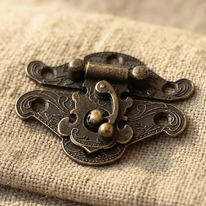 May include: A bronze-toned metal box latch featuring an ornate, embossed scrollwork pattern. The hardware consists of two interlocking pieces with four circular screw holes for mounting, a central swivel hook mechanism, and a hinged top section.