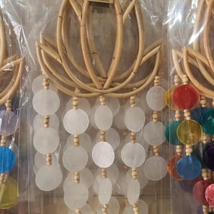 Puede incluir: Atrapasueños con diseño de flor de loto hecho de bambú. El atrapasueños tiene una serie de discos redondos blancos con forma de concha colgando, intercalados con cuentas de madera. El atrapasueños está colgado de una rejilla metálica.