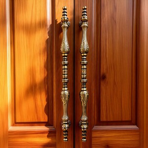 Puede incluir: Dos manijas de puerta ornamentadas de estilo antiguo con acabado de latón están montadas en una puerta de madera. Las manijas tienen un diseño vertical con detalles intrincados y están colocadas simétricamente, agregando elegancia a la entrada.