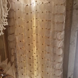 Può includere: Una tenda di perline composta da dischi rotondi e traslucidi di conchiglia bianco sporco. I dischi sono collegati con piccole perline marrone scuro. La tenda è appesa verticalmente, creando un effetto decorativo e strutturato. Il tema generale è bohémien e naturale.