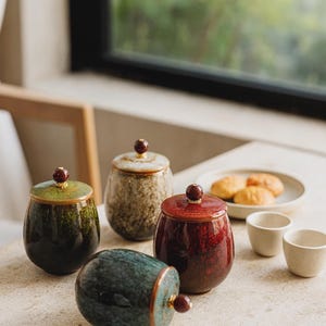Puede incluir: Conjunto de tarros de cerámica con tapas, en tonos verdes, marrones y rojos. Cada tarro tiene un pequeño pomo redondo en la parte superior. También se ven dos tazas blancas pequeñas y un plato con pasteles.