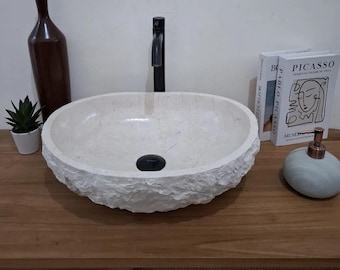 Puede incluir: Un lavabo de piedra de color crema, de forma ovalada, con un exterior rugoso y texturizado. Se instala un grifo y un desagüe negros. El lavabo se encuentra sobre un mueble de madera, con objetos decorativos.