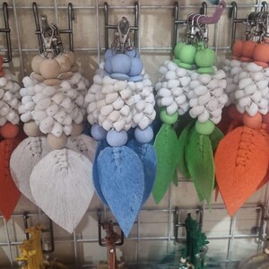 Pode incluir: Uma coleção de chaveiros coloridos com um design floral. Cada chaveiro apresenta um grupo de elementos brancos em forma de contas, encimados por esferas coloridas e detalhes em forma de folha em várias tonalidades, incluindo verde, laranja, azul e branco. Eles penduram de clipes de metal prateados.