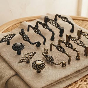 May include: A collection of metal cabinet hardware including various drawer pulls and knobs. The pieces feature dark bronze and antique brass finishes with intricate twisted wire cage designs and solid circular bases, arranged in a group.