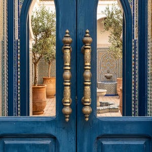 May include: A pair of blue wooden doors with ornate brass handles. The doors open to a courtyard with olive trees in terracotta pots and a tiled wall with a fountain. The door frame has decorative blue and white tile accents.