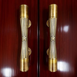May include: A pair of brass door handles with a star motif. The handles are cylindrical, featuring vertical grooves and a star emblem. They are mounted on a dark red wooden door.
