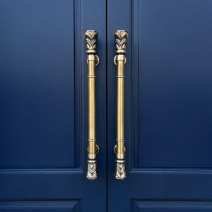 May include: Two brass door handles with decorative floral detailing, mounted on a deep blue door. The handles are vertical, with a cylindrical body. The door has a paneled design. The handles are likely around 100 cm in length.