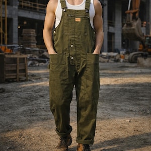 Op de afbeelding: Olijfgroene werkoverall met meerdere zakken en knoopsluitingen. De overall heeft verstelbare bandjes en een kleine logopatch op de borst. De persoon draagt een witte tanktop en bruine werkschoenen. De achtergrond toont een bouwplaats.