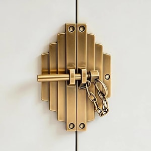 May include: A brass door bolt latch featuring a stepped, geometric plate design with vertical ridges. The hardware includes a horizontal sliding bolt and a short security chain attached to the side, secured with visible screws on a white surface.