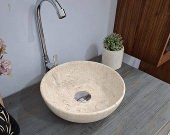 Puede incluir: Un lavabo redondo de mármol beige con un grifo cromado. El lavabo tiene un desagüe central y está sobre una superficie de madera gris. Una pequeña planta en maceta es visible en el fondo.