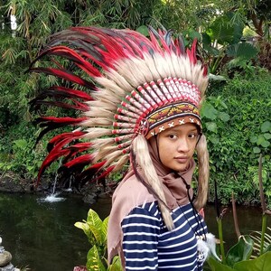 May include: A Native American headdress with red, black, and white feathers. The headdress features a beaded band with geometric patterns. The person in the image is wearing a striped shirt and a brown head covering.