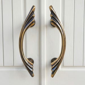 May include: Pair of bronze-colored door handles with a curved, Art Deco-inspired design. The handles are mounted on a white paneled door. The handles have a decorative, fan-like shape at the top and bottom.